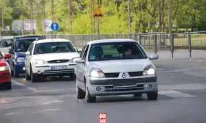 Samochody na drodze, w tym srebrny Renault Clio i biały Ford, zbliżają się do przejścia dla pieszych. Brak tablic rejestracyjnych na niektórych pojazdach.