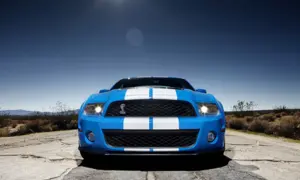 Niebieski Shelby GT500 z białymi pasami na masce i dachu, zaparkowany na pustynnej drodze pod błękitnym niebem.