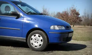 Błękitny Fiat Albea stoi na polnej drodze, w tle drzewa i trawa.