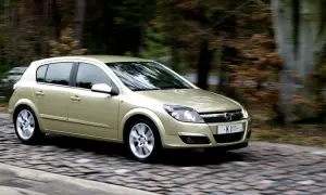 Złoty Opel Astra H 1.7 CDTI w ruchu na brukowanej drodze, otoczony rozmazanym tłem lasu.