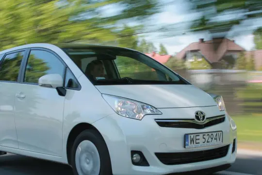 Biała Toyota Verso S w ruchu, z rozmytym tłem zieleni i domów.