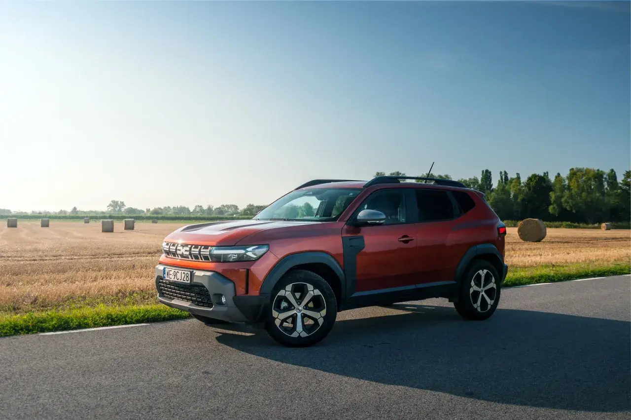 Nowa Dacia Duster w kolorze rudym, zaparkowana na poboczu drogi, z polem słomy w tle.