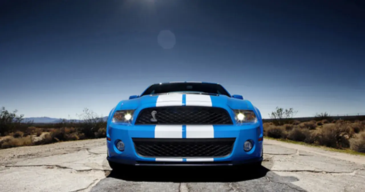 Niebieski Shelby GT500 z białymi pasami na masce i dachu, zaparkowany na pustynnej drodze pod błękitnym niebem.