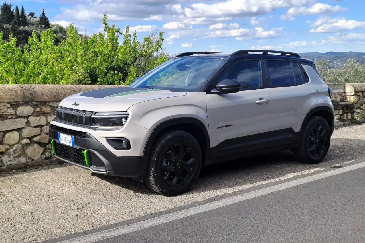 Jasnoszary Jeep Avenger 4xe z zielonymi akcentami na masce i zderzaku, zaparkowany na tle kamiennego muru i zieleni.
