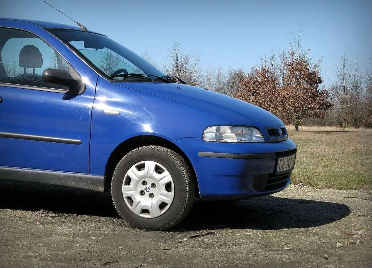 Błękitny Fiat Albea stoi na polnej drodze, w tle drzewa i trawa.