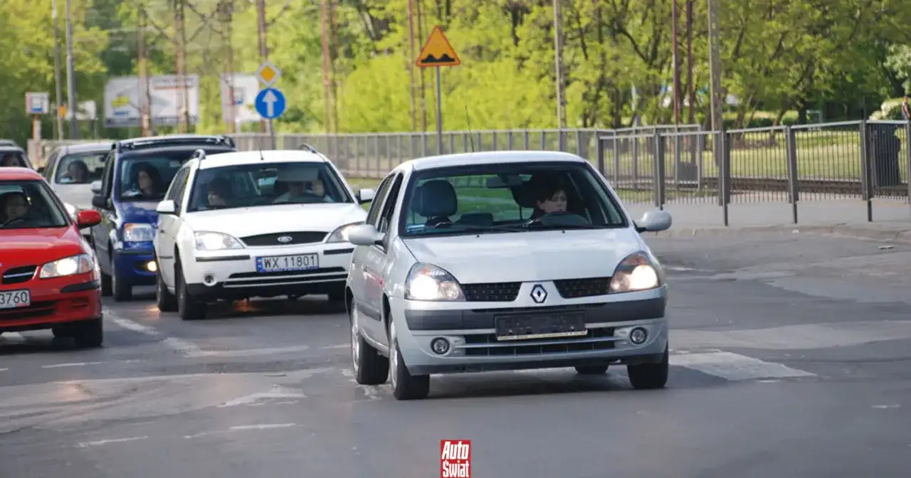 Samochody na drodze, w tym srebrny Renault Clio i biały Ford, zbliżają się do przejścia dla pieszych. Brak tablic rejestracyjnych na niektórych pojazdach.