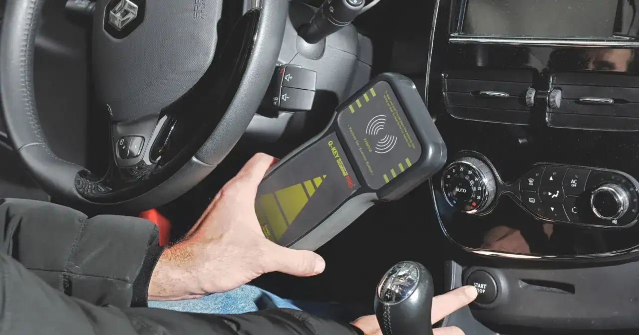 Dłoń trzyma skaner "Q-KEY Scanner PRO" do systemu keyless, obok kierownicy i przycisku start w samochodzie.