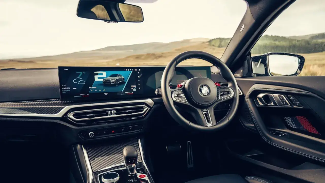 Wnętrze BMW M2 G87 z dużym ekranem, kierownicą M i sportową dźwignią biegów. Widok na krajobraz za oknem.