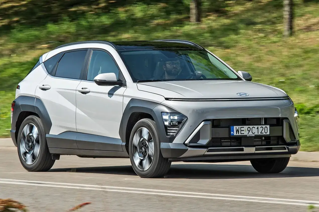 Nowy Hyundai Kona 2025 w kolorze białym z czarnym dachem, jadący po drodze wśród zieleni.