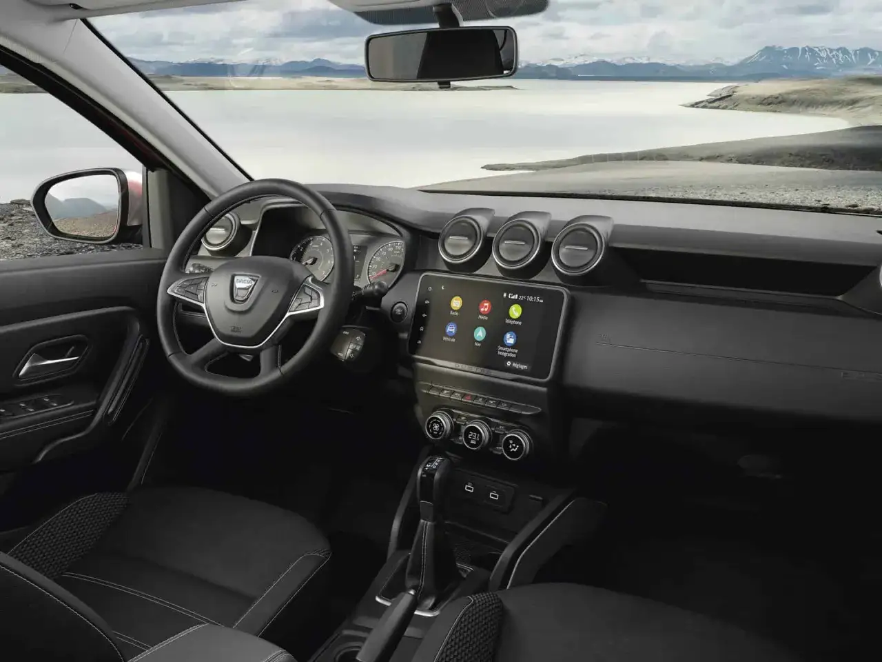 Wnętrze nowej Dacii Duster z ekranem dotykowym i widokiem na jezioro i góry.
