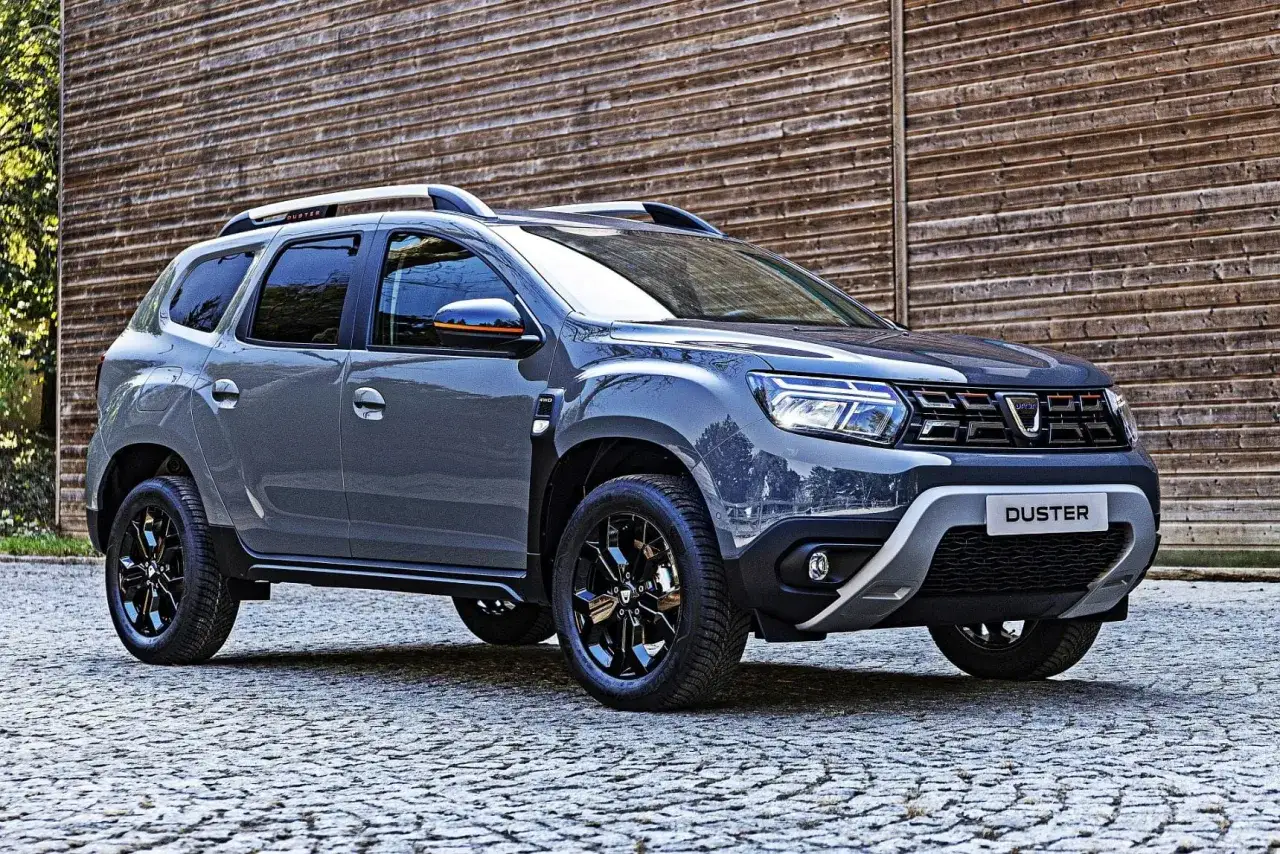 Szary Dacia Duster 2026 z czarnymi felgami i pomarańczowymi akcentami, zaparkowany na bruku przed drewnianą ścianą.