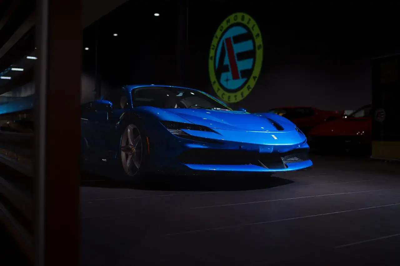 Błękitne Ferrari SF90 błyszczy w ciemnym salonie, obok innych sportowych samochodów.