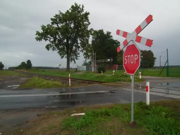 Przejazd kolejowy z zaporami i znakiem STOP. Torowisko przecina mokrą drogę, a w tle widać drzewa i boisko.