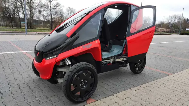 Czerwony, trójkołowy polski samochód elektryczny Trigga z otwartymi drzwiami, zaparkowany na placu.