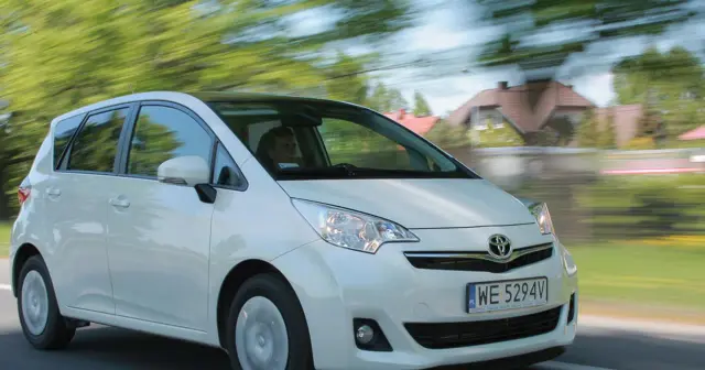 Biała Toyota Verso S w ruchu, z rozmytym tłem zieleni i domów.