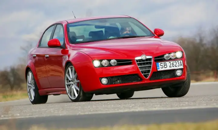 Czerwone Alfa Romeo 159 1.9 JTD na drodze, z kierowcą za kierownicą.
